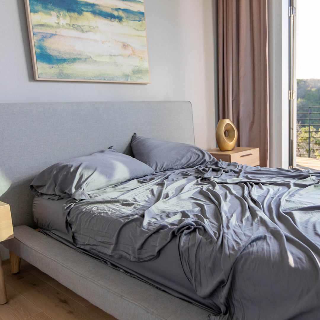 Gray bedding on a bed in a bedroom with a painting and window in the background.