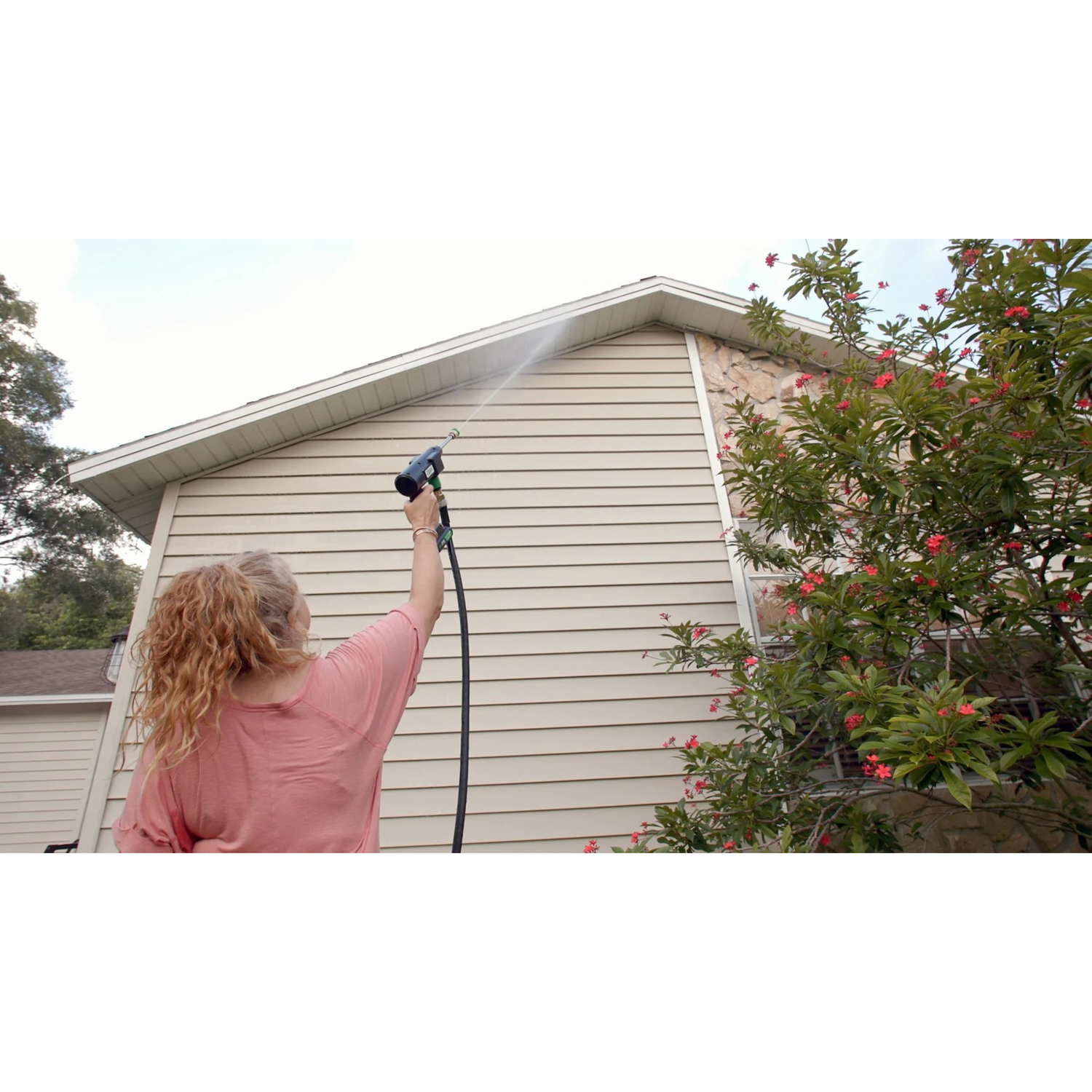 Person using a pressure washer on a house exterior with trees and flowers in the background.