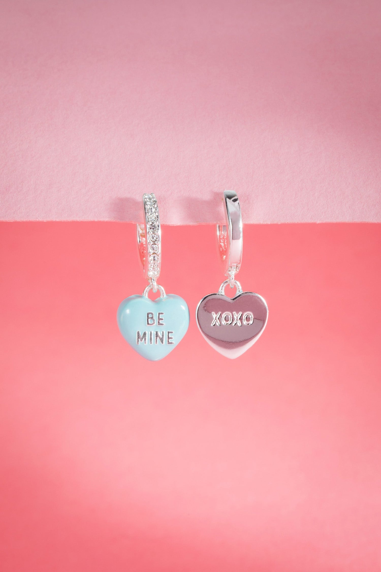 Heart-shaped earrings with 'Be Mine' and 'XOXO' charms on a pink background