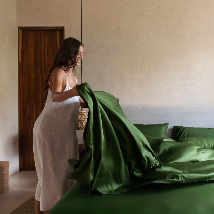 Woman in a white dress arranging green satin sheets on a bed in a bedroom.