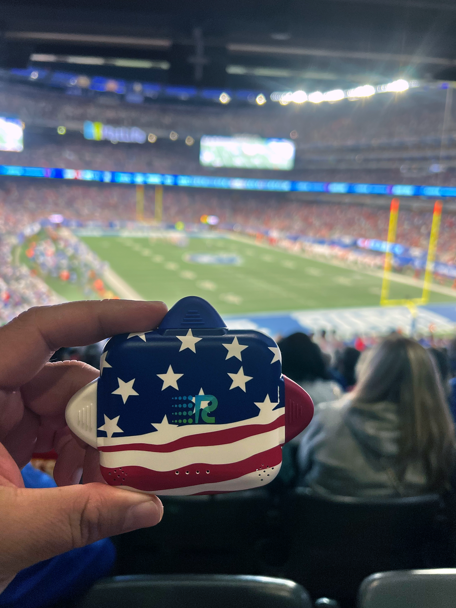 Hand holding a small American flag-themed object with a football stadium in the background