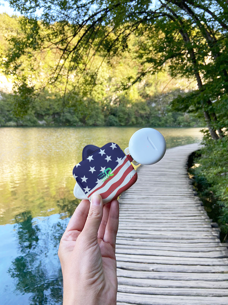 Hand holding a decorative object with an American flag design on a wooden bridge over a lake.