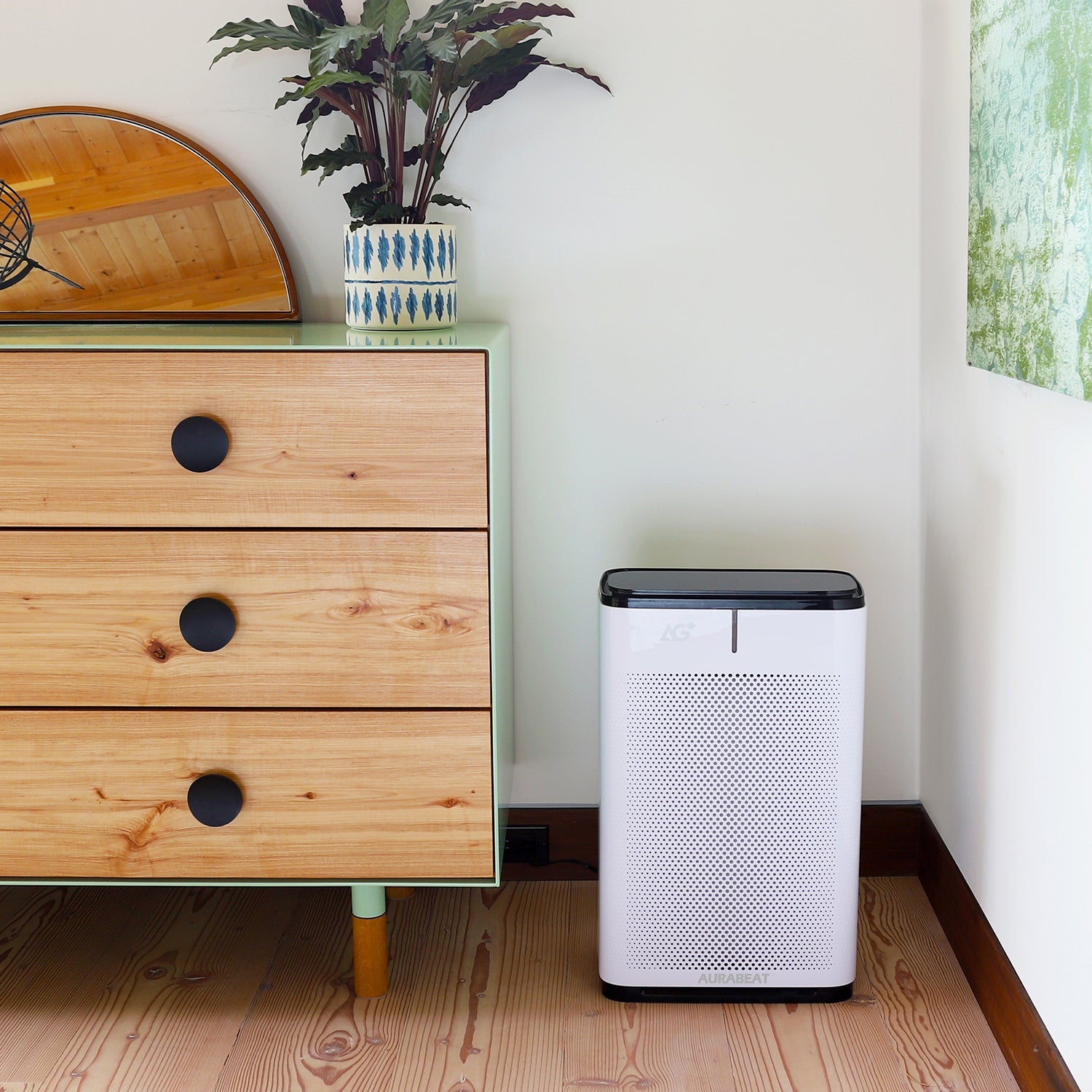 White air purifier on a wooden floor next to a wooden dresser with a plant on top.