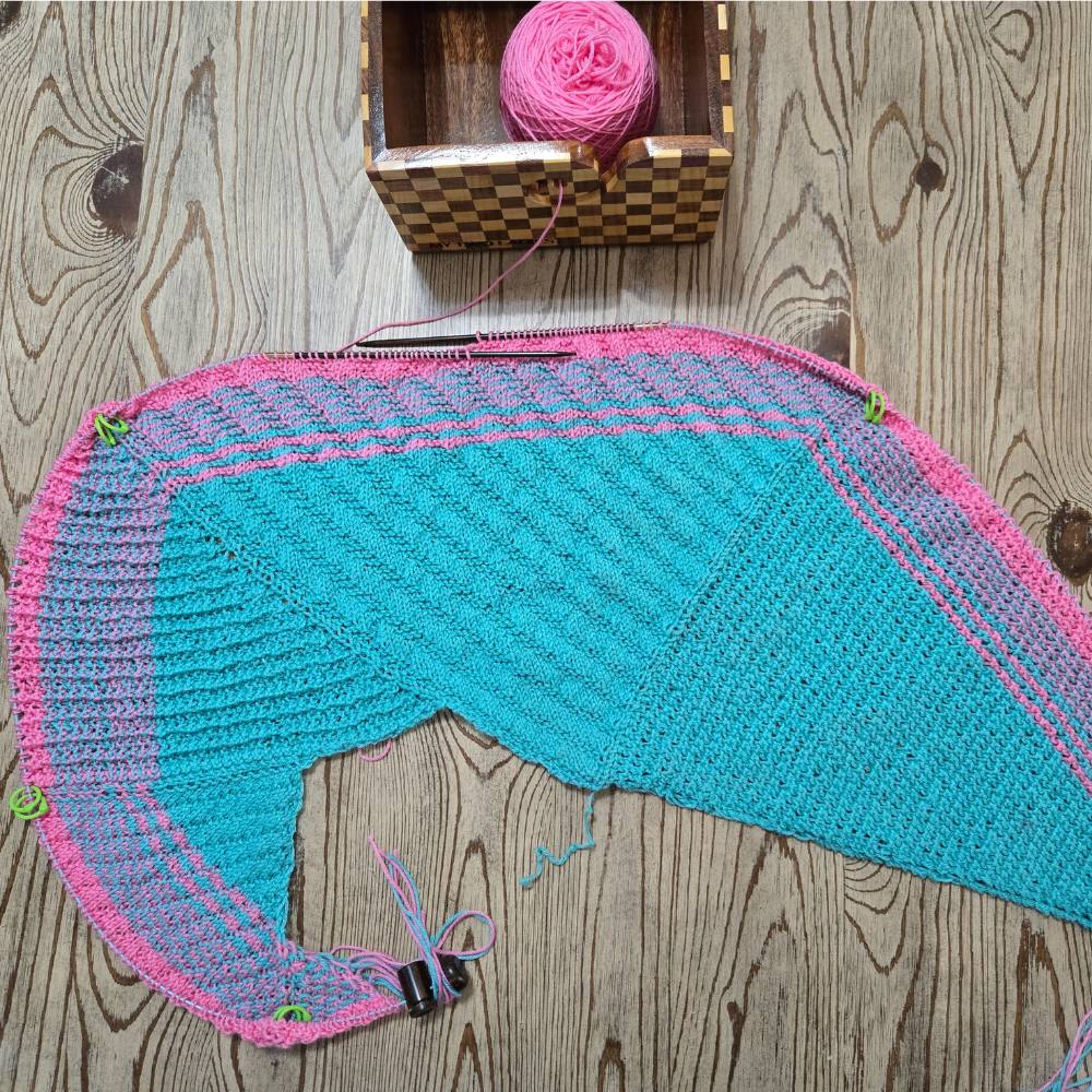 Knitted fabric with pink and blue pattern on a wooden surface with yarn and knitting tools.