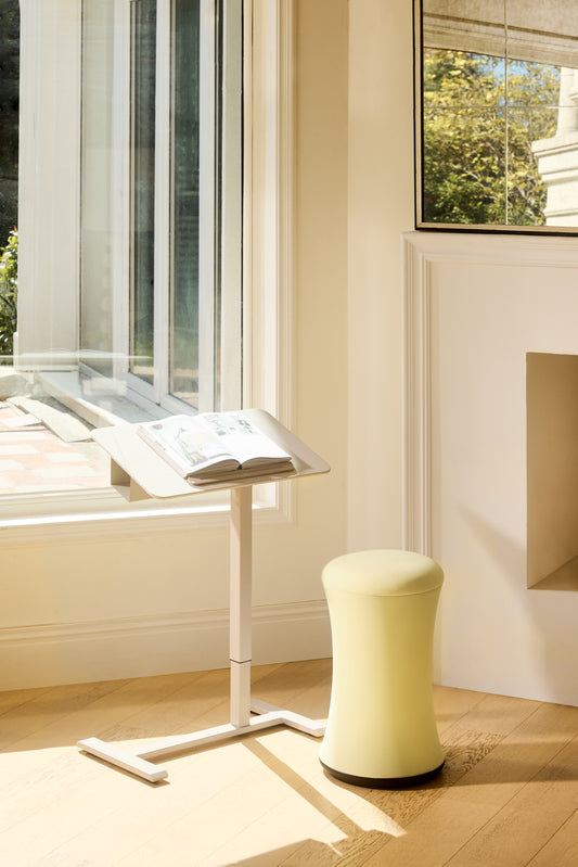 Light-colored stool next to a window with a book on a stand.