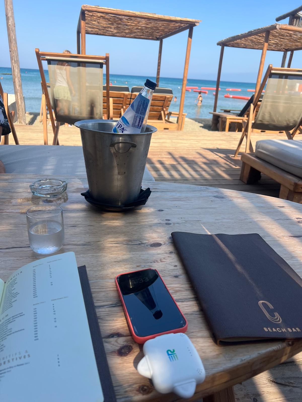 A beachside wooden table at a resort with a metal ice bucket holding a water bottle, a glass, an open menu, a red phone case, a black Beach Bar menu cover, and a small white device. Lounge chairs, umbrellas, and the ocean are visible in the background.