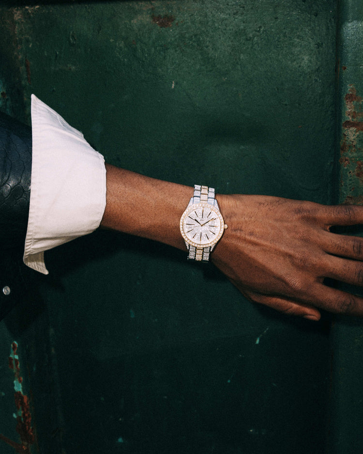 A close-up of a wrist wearing the JBW Cristal 39 | J6346D luxury diamond watch with Swiss movement. The person, dressed in a white shirt and dark blazer, rests their hand on a textured dark green surface.