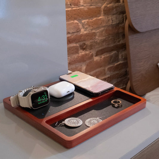 A wireless charging pad with integrated Apple Watch charger, featuring a black and oak design, with a smartphone, smartwatch, and earbuds being charged.