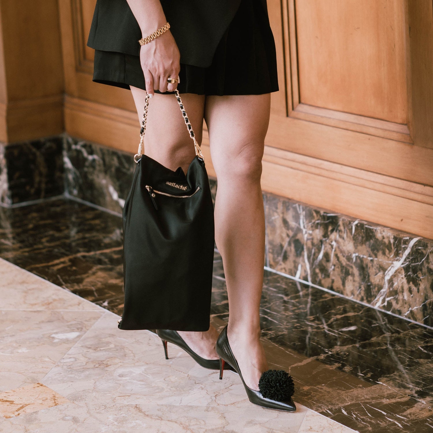 Person holding a black handbag with a gold chain strap, wearing high-heeled shoes, in an indoor setting.
