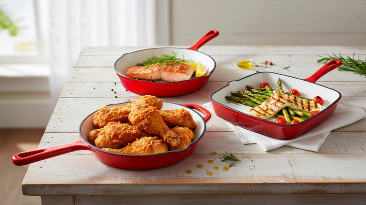 Red frying pans with fried chicken, vegetables, and salmon on a wooden table.
