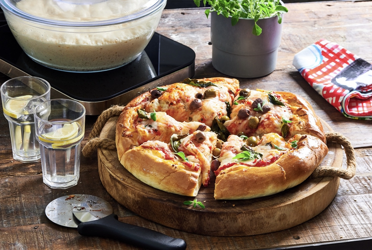 Pizza on a wooden board with glasses of water and a plant in the background