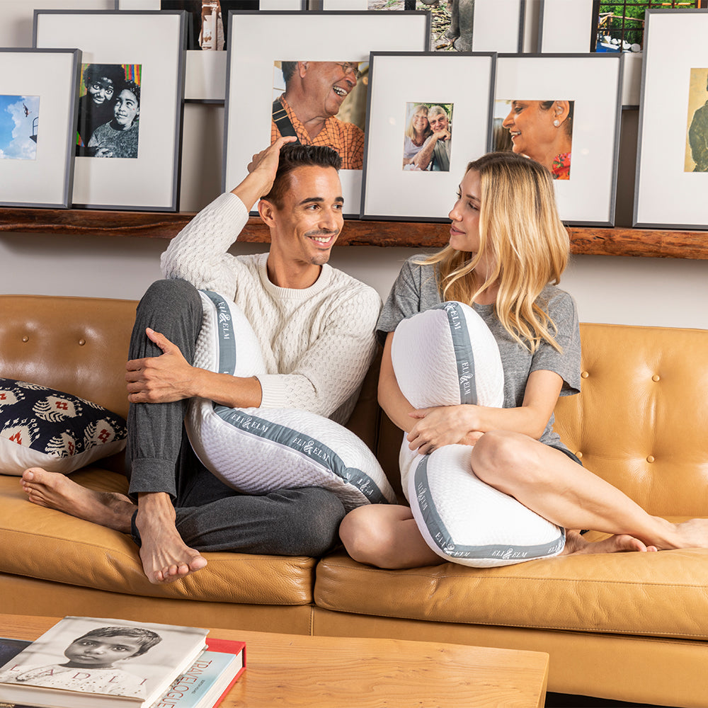 Man and woman sitting on a couch with baby bump pillows, surrounded by framed photos.