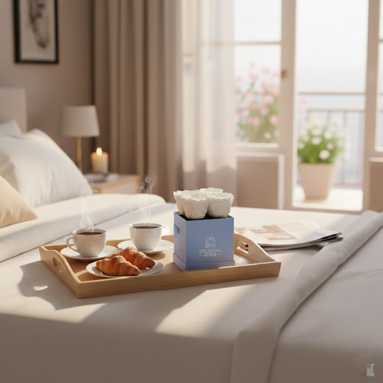 Breakfast tray with coffee, croissants, and flowers on a bed in a bright bedroom.
