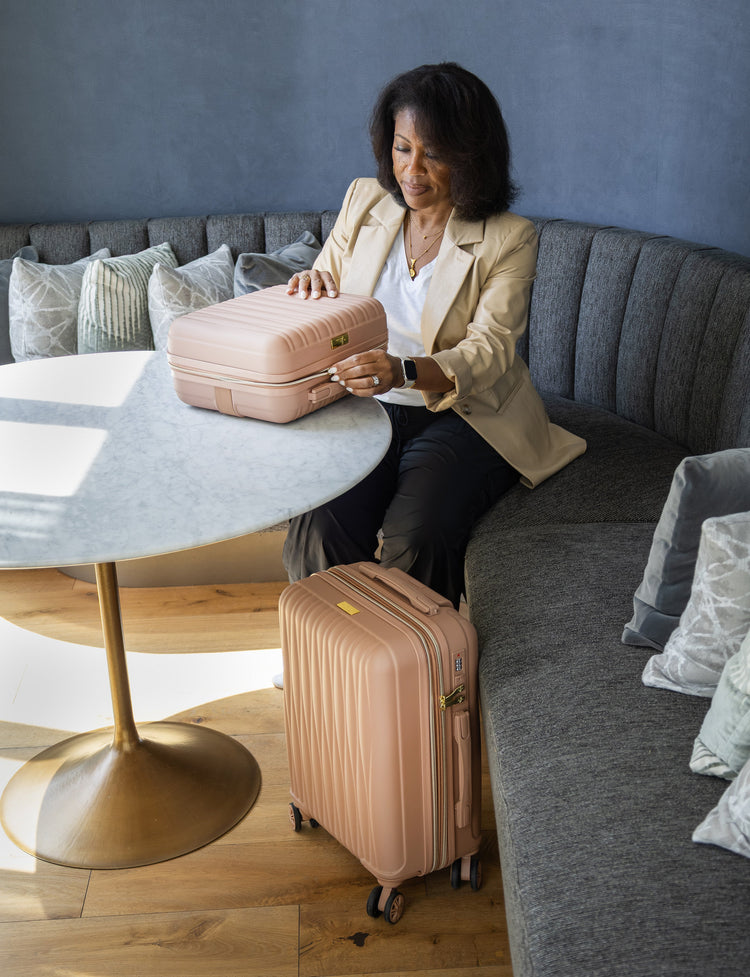 Woman sitting on a couch with a pink suitcase and travel case in a living room setting.
