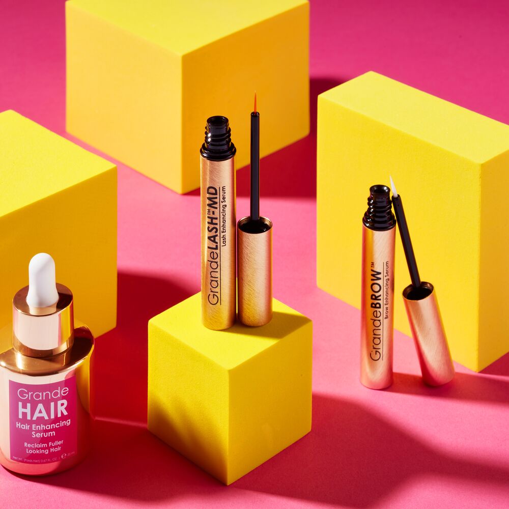 Makeup products including a serum and two mascara tubes on yellow and pink geometric blocks.