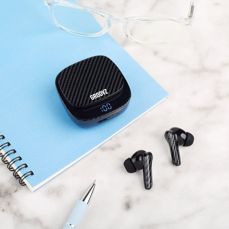 Black wireless earbuds with charging case on a marble surface with glasses and a pen.