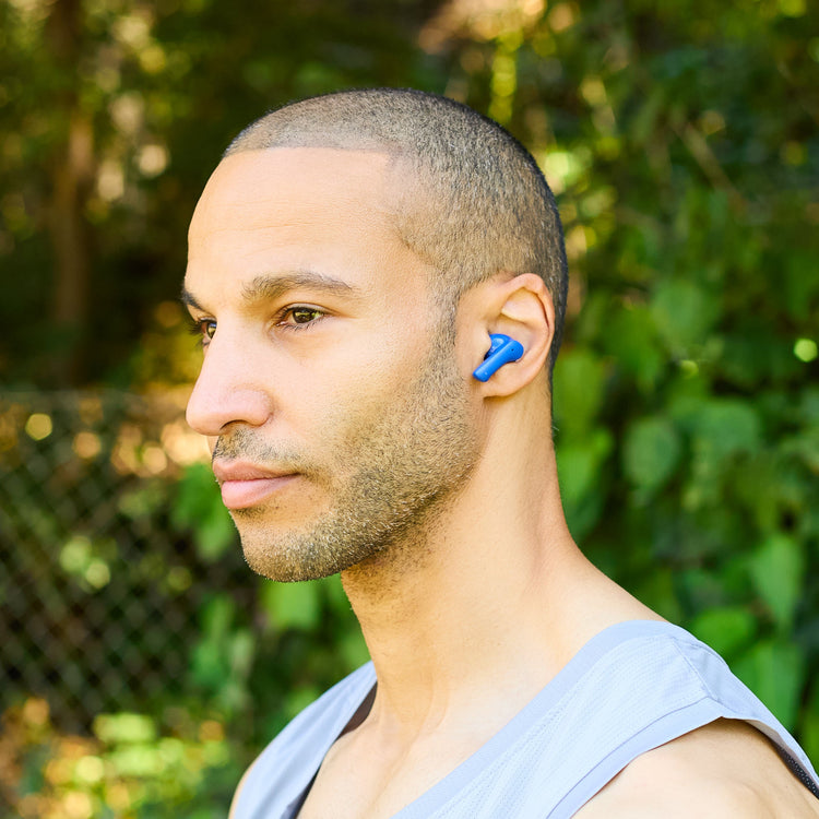 Man wearing a blue earbud outdoors with greenery in the background