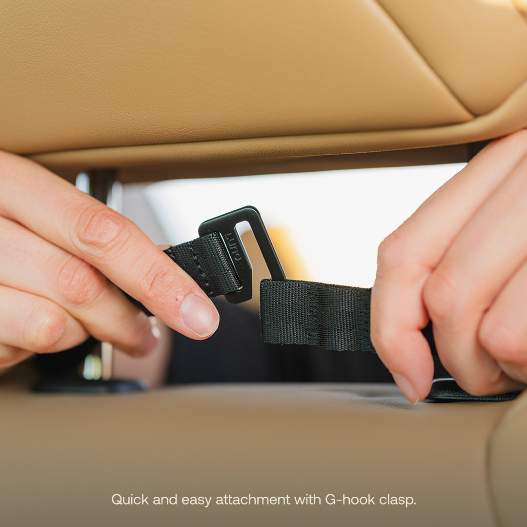 Close-up of hands fastening a black strap with a G-hook clasp on a tan car seat, showcasing the Luno® Seatback Organizer. The image text reads: "Quick and easy attachment with G-hook clasp for seamless vehicle organization or essential car camping setup.