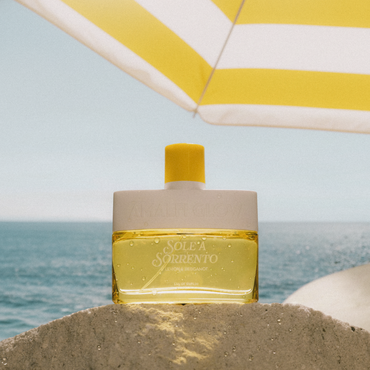 Solea Sorrento perfume bottle on sand with ocean and yellow umbrella in the background
