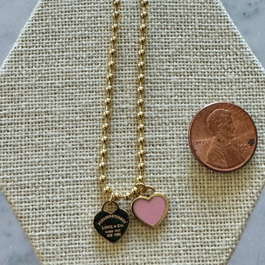 Gold necklace with heart-shaped pink pendant on a textured surface with a penny for scale.
