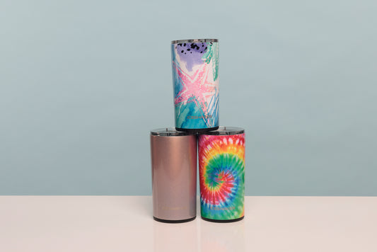 Three insulated tumblers stacked in a pyramid on a white surface against a light blue-gray background. The bottom-left tumbler is metallic rose gold, the bottom-right tumbler has a rainbow tie-dye spiral, and the top tumbler has a pastel multicolor design with a pink starfish.