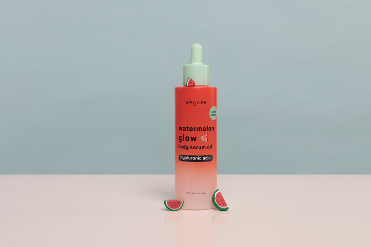 A bottle of SPALiPS Watermelon Glow Daily Serum Oil with a pink-to-white gradient body, a mint green dropper cap, and watermelon slice props on a light pink surface against a gray background.