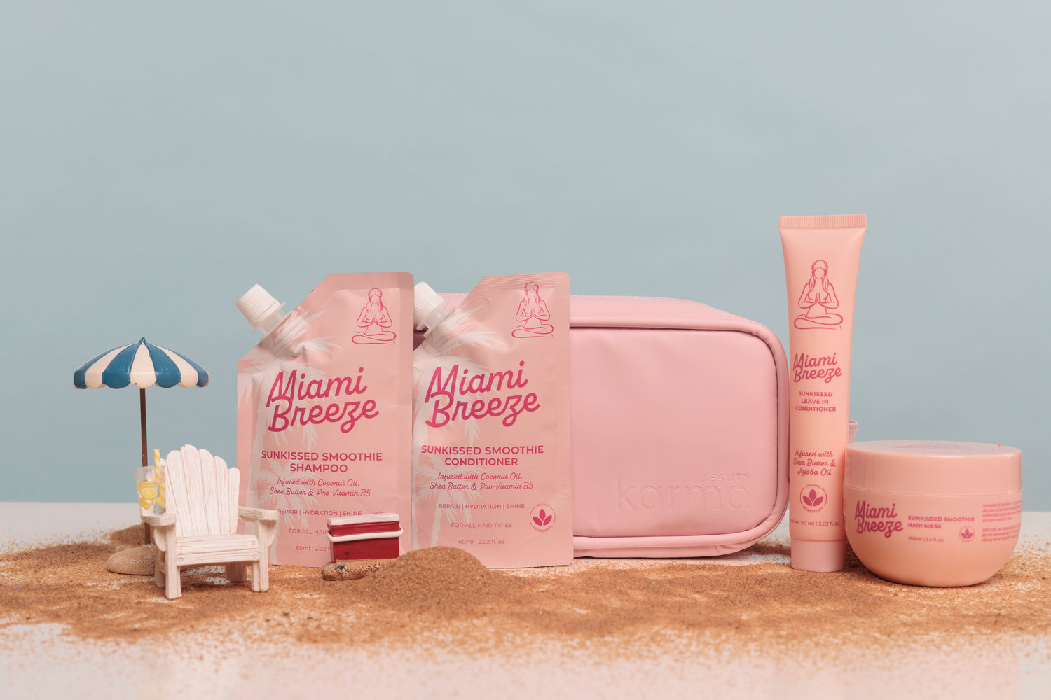 Miami Breeze skincare set in soft pink packaging, including a pouch of smoothie shampoo, a tube of smoothie conditioner, a round jar, and a zip pouch, arranged on a sandy surface with a small beach chair and umbrella prop against a pale blue background.