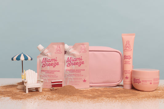 Miami Breeze skincare set in soft pink packaging, including a pouch of smoothie shampoo, a tube of smoothie conditioner, a round jar, and a zip pouch, arranged on a sandy surface with a small beach chair and umbrella prop against a pale blue background.