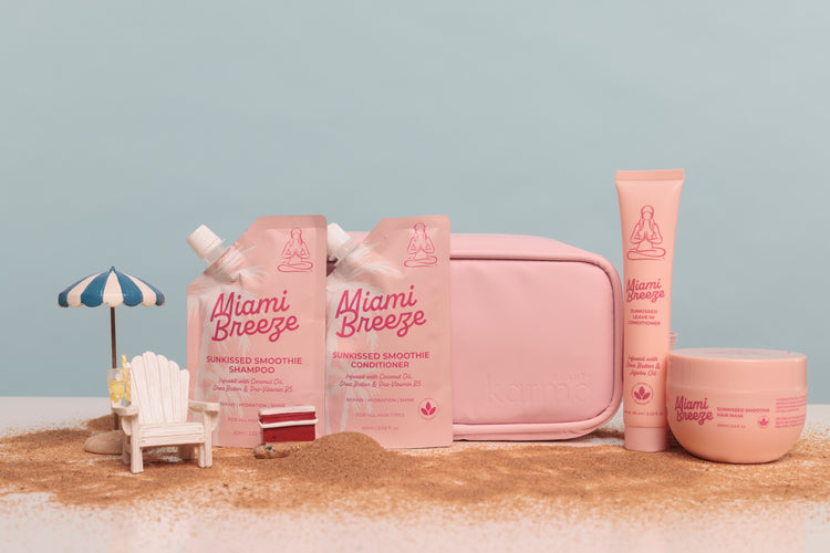 Miami Breeze skincare set in soft pink packaging, including a pouch of smoothie shampoo, a tube of smoothie conditioner, a round jar, and a zip pouch, arranged on a sandy surface with a small beach chair and umbrella prop against a pale blue background.