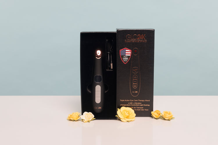 A black skincare device displayed inside its open black retail box next to the branded outer box, with a few yellow flowers placed on the surface below. The background is a clean light blue wall and white tabletop.