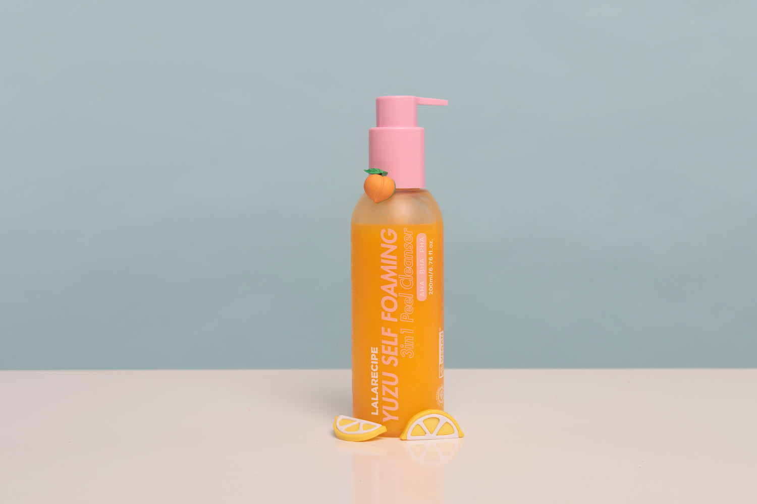 A bottle of JalaRubic Yuzu Self Foaming body wash with a pink pump dispenser, set on a white surface with yellow lemon slice props against a light blue-gray background.