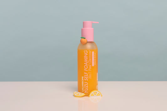 A bottle of JalaRubic Yuzu Self Foaming body wash with a pink pump dispenser, set on a white surface with yellow lemon slice props against a light blue-gray background.