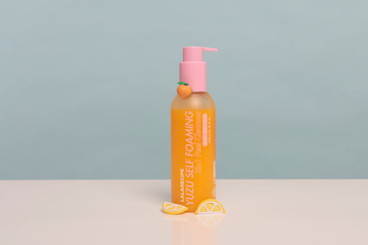 A bottle of JalaRubic Yuzu Self Foaming body wash with a pink pump dispenser, set on a white surface with yellow lemon slice props against a light blue-gray background.