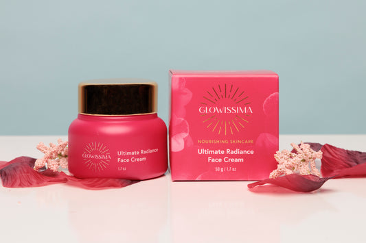 Glowissima Ultimate Radiance Face Cream in a pink jar with a gold lid, shown next to its matching pink box on a white surface with dried rose petals against a light blue background.