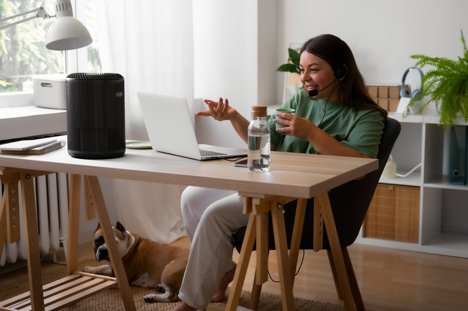 Person sitting at a desk with a laptop, drinking from a cup, and a dog lying on the floor.