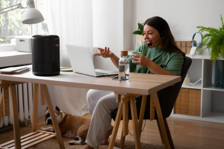 Person sitting at a desk with a laptop, drinking from a cup, and a dog lying on the floor.