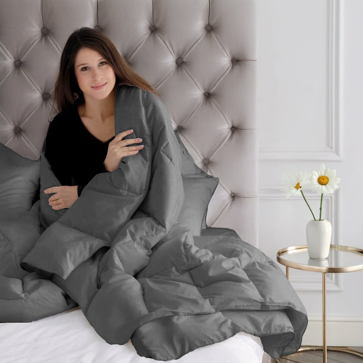 Woman wrapped in a gray comforter sitting on a tufted headboard in a bedroom.