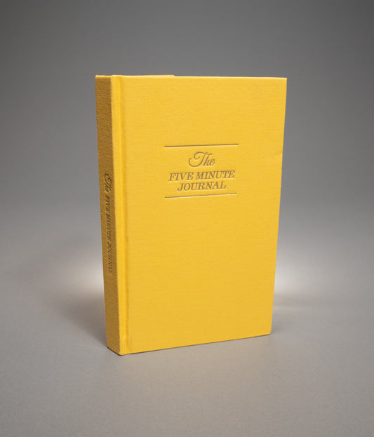 Yellow journal titled 'The Five Minute Journal' on a gray background