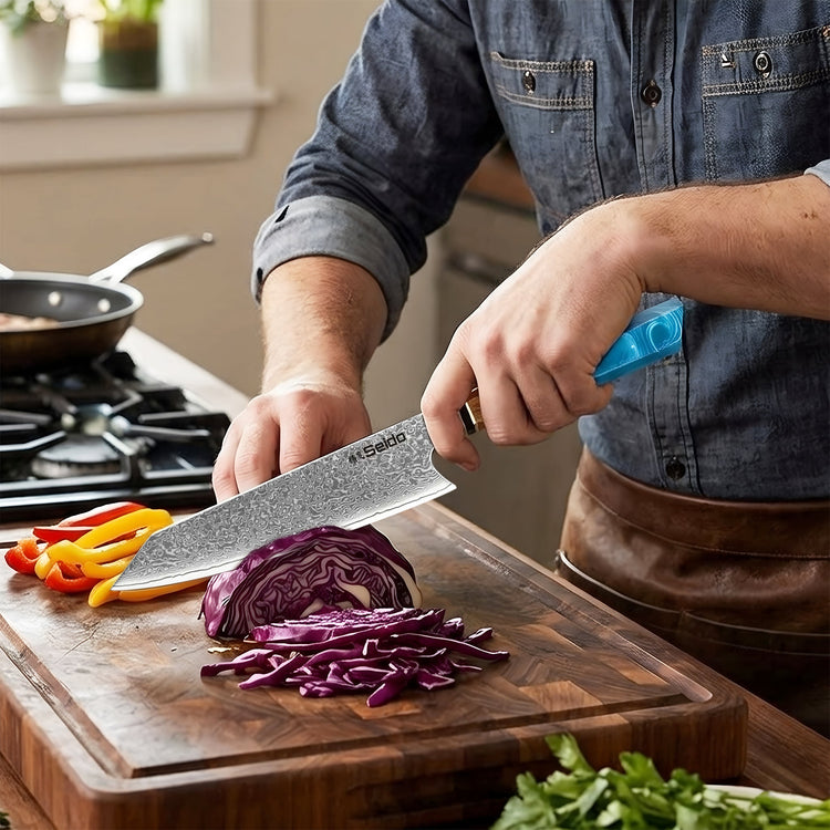 A person in a kitchen wearing a denim shirt and brown apron slices purple cabbage on a wooden cutting board with a large Santoku knife with a textured gray blade and a bright blue handle.