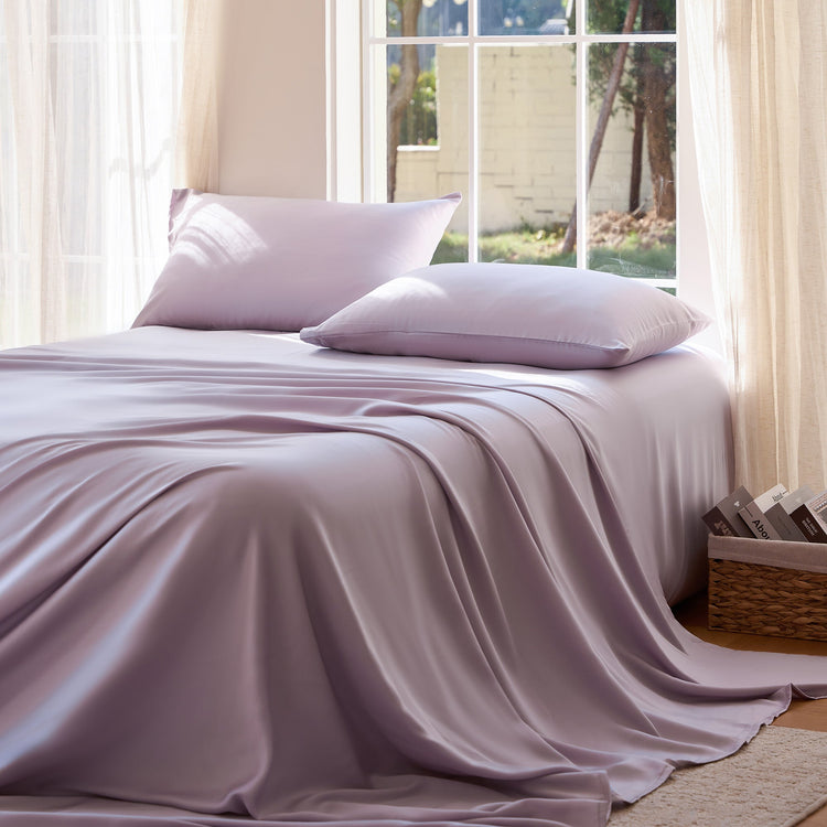 Bed with light purple bedding and pillows in a bright room with large windows.