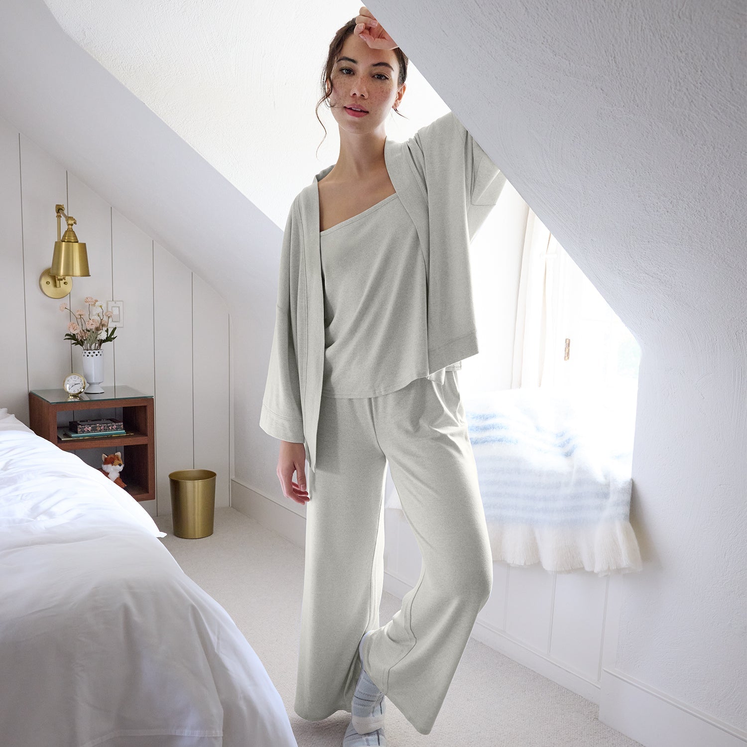 Woman in a light gray outfit standing in a bright bedroom.