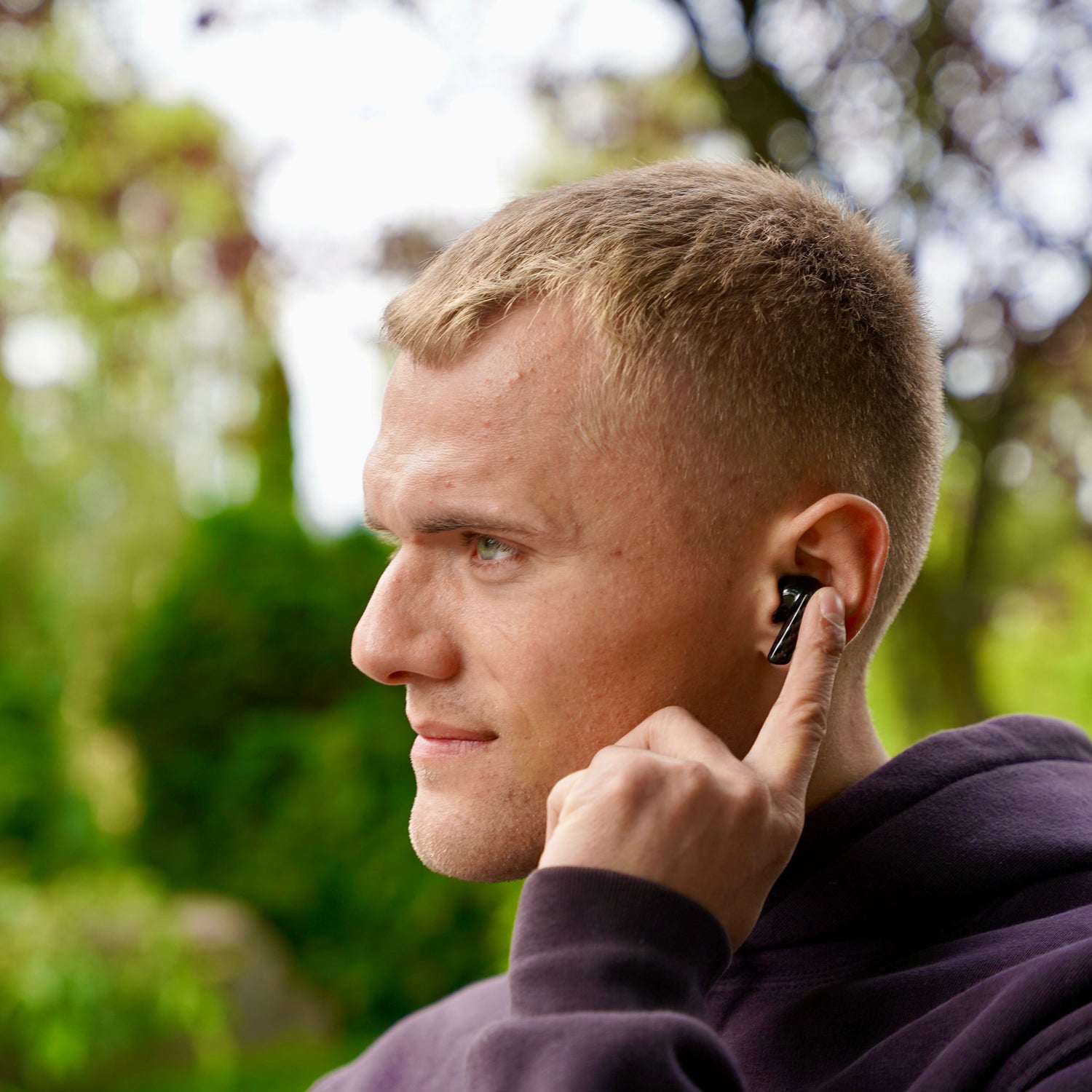 Man with short hair holding a phone to his ear outdoors with greenery in the background