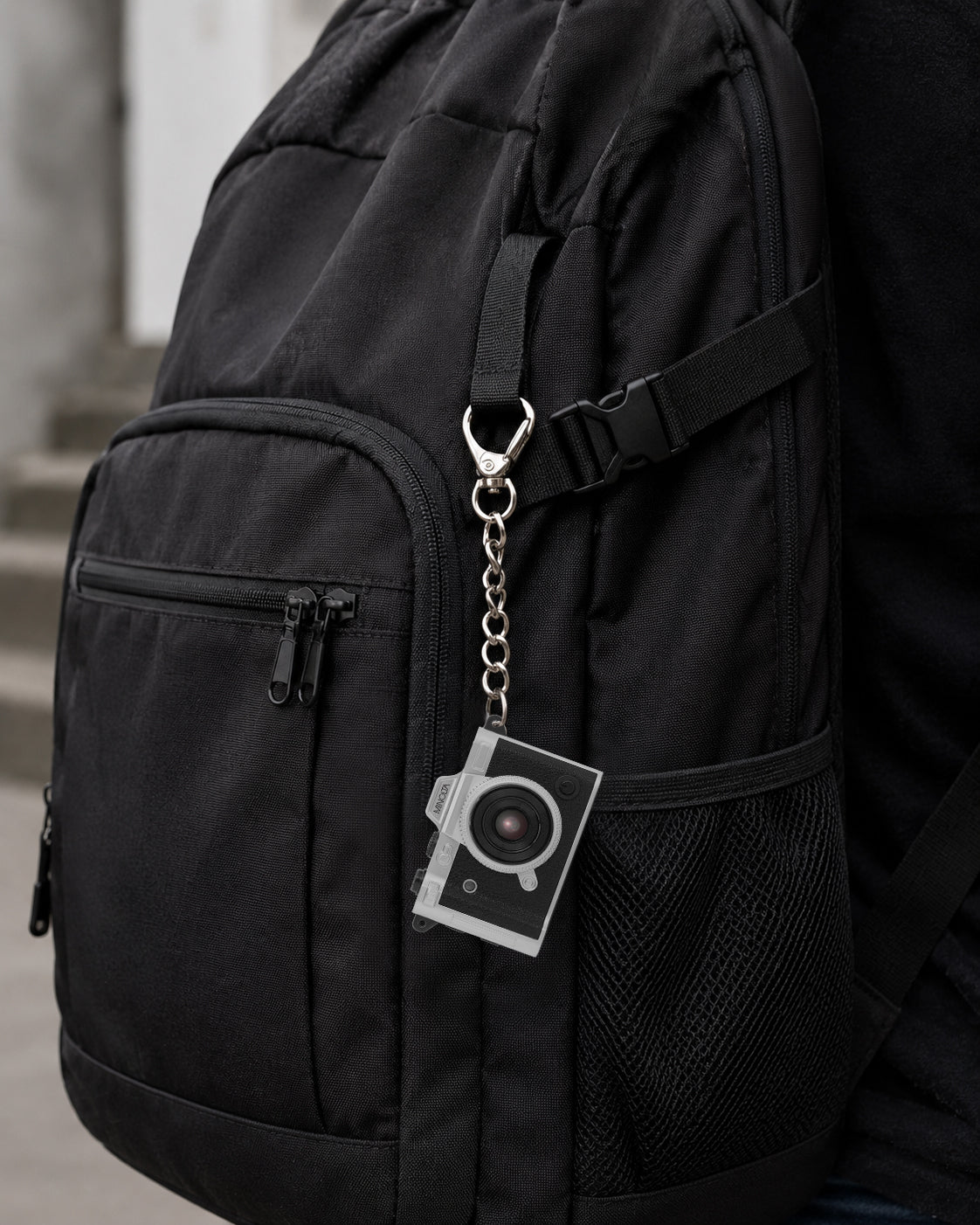 A close-up of a black backpack with a silver camera-shaped keychain attached to a strap, shown against a blurred outdoor background.