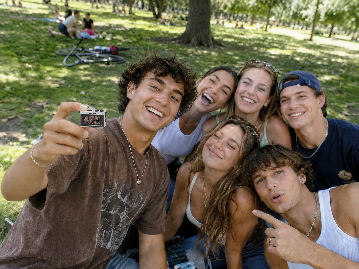 A group of five young friends smiling and taking a selfie in a sunny park with trees and grass in the background.