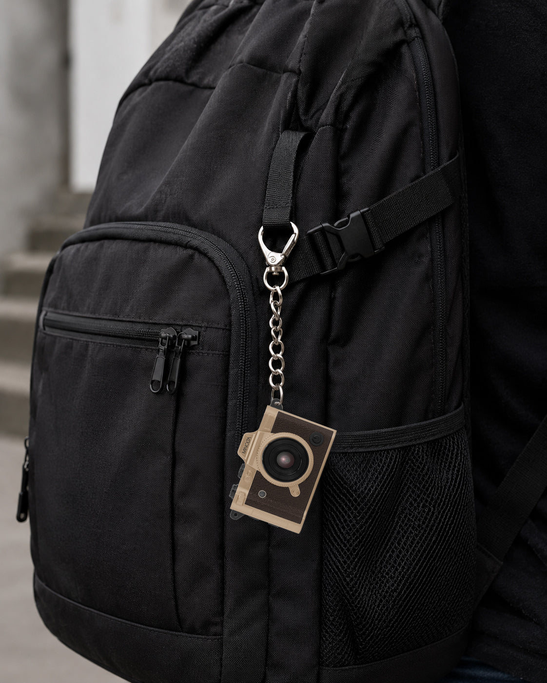 A close-up of a black backpack with a side pocket, mesh pocket, and a silver chain keychain holding a small rectangular camera-shaped accessory, shown against a blurred outdoor background.