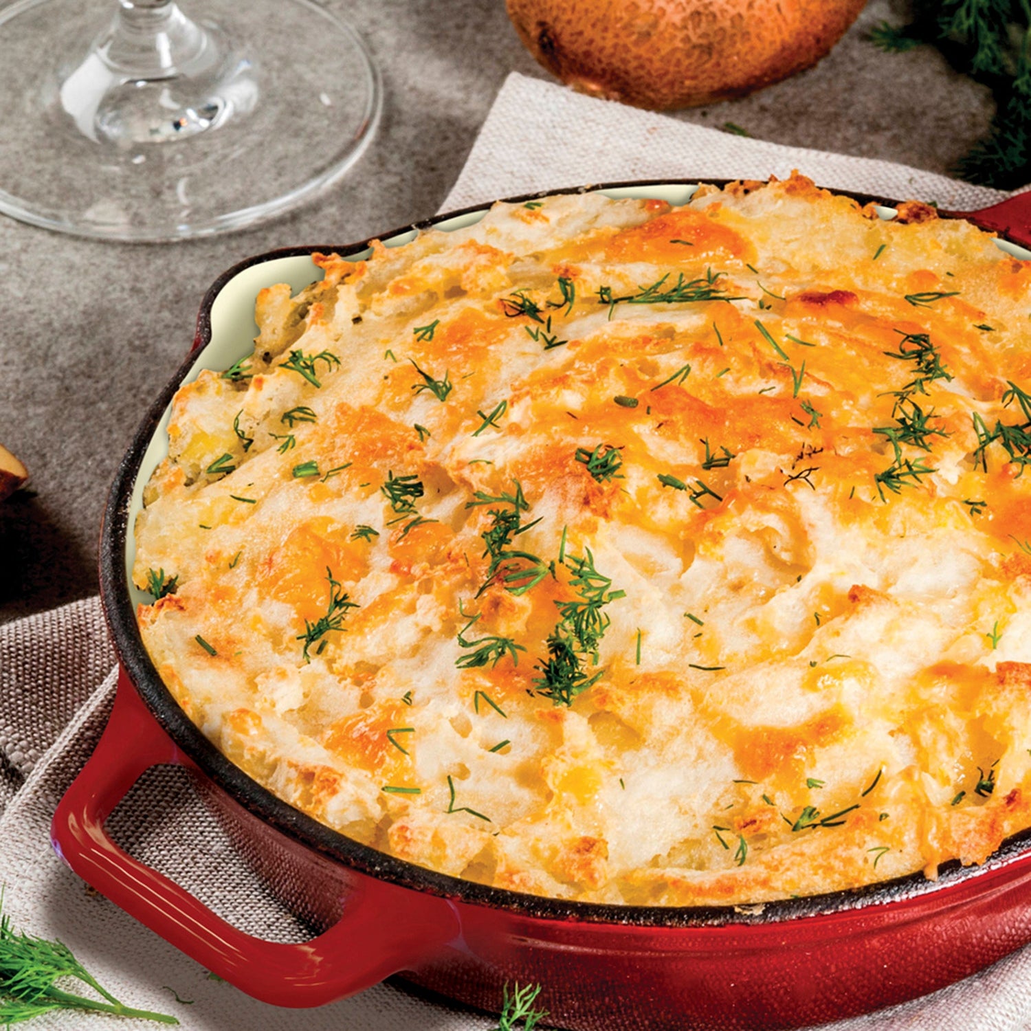 Cheesy dish in a red skillet on a table with a glass and onion in the background