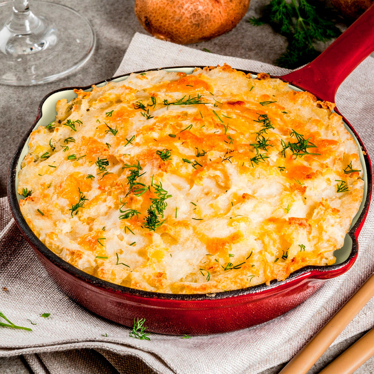 Cheesy dish in a red skillet on a textured surface with a glass and onion in the background.