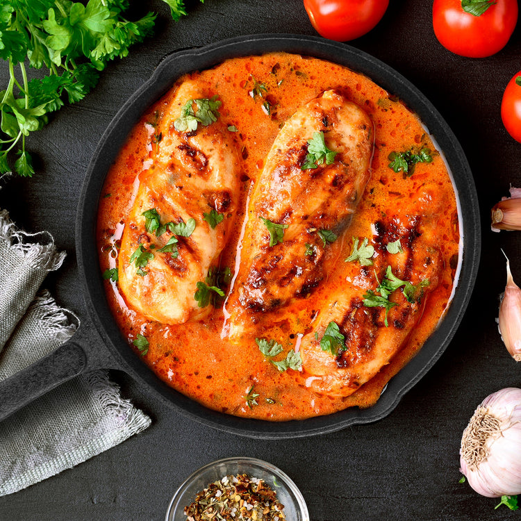 Chicken dish with red sauce in a black skillet on a dark surface with tomatoes and herbs.