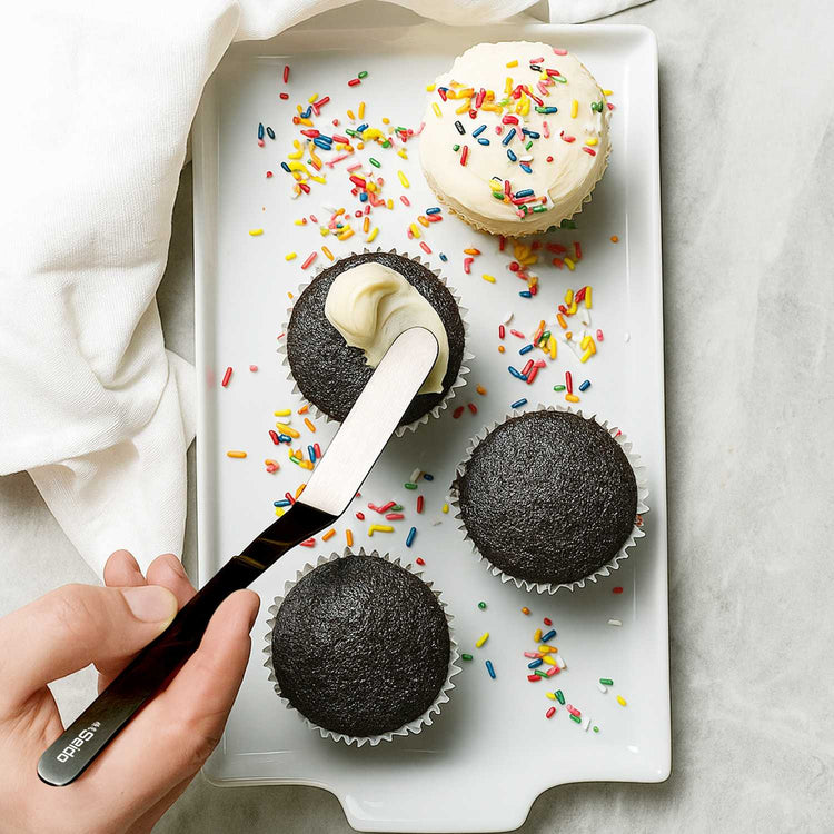 Person frosting black cupcakes with colorful sprinkles on a white tray.