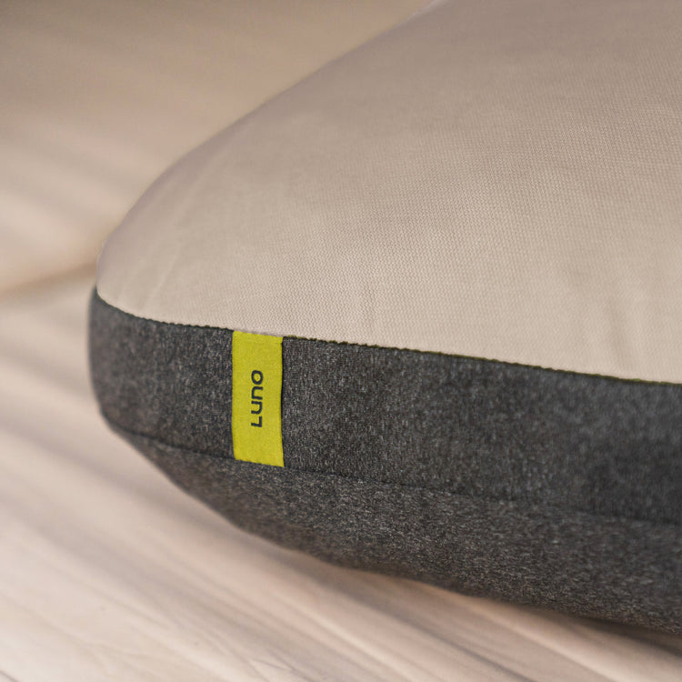 Close-up of the Luno® Packable Camping Pillow, featuring a beige top, dark gray bottom, and a small yellow Luno® tag, displayed on a wooden surface.